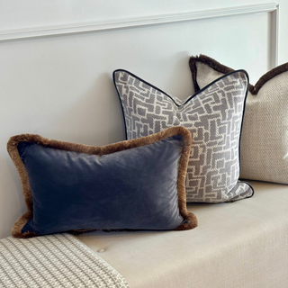 Three decorative throw pillows arranged on a cream upholstered bench in a real interior setting with a staircase visible in the background. The pillows include a navy velvet pillow with brown faux-fur trim, a light gray and white geometric pattern pillow with dark trim, and a textured beige pillow with dark brown trim.