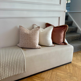 A beige upholstered bench or ottoman in a styled interior, decorated with three decorative throw pillows in beige, cream, and rust brown, with a textured cream throw blanket draped over the left side.