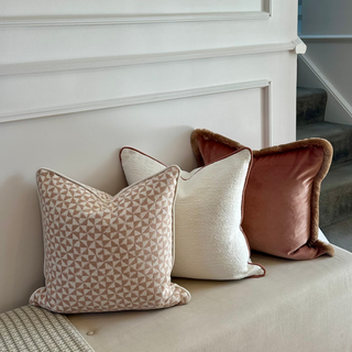 Three decorative throw pillows in neutral and muted tones arranged on a beige upholstered bench against a white paneled wall, with a framed artwork above and stairs visible in the background.