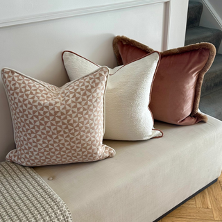 Three decorative throw pillows in neutral and muted tones arranged on a beige upholstered bench in a home interior setting.