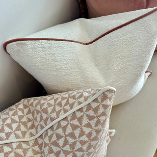 A styled collection of home decor throw pillows, featuring a prominent cream bouclé pillow with rust-brown piping, a dusty pink velvet pillow with a brown faux-fur trim, and a beige and cream geometric patterned pillow.