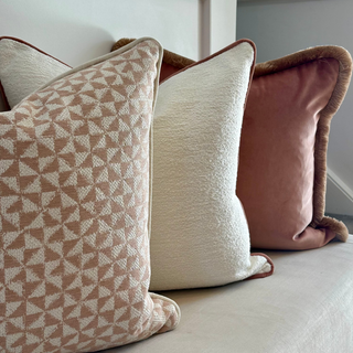Three decorative throw pillows arranged on a light beige sofa in front of a white paneled headboard. The left pillow has a beige and off-white geometric triangle pattern, the center pillow is cream with a soft textured surface, and the right pillow is dusty rose pink with a dark brown faux-fur trim.