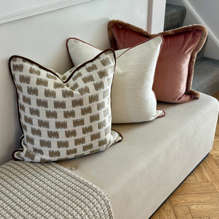 Three decorative throw pillows arranged on a light beige upholstered bench in a home interior. The pillows include a cream pillow with a brown geometric pattern, a solid cream pillow, and a dusty rose velvet pillow with brown trim.