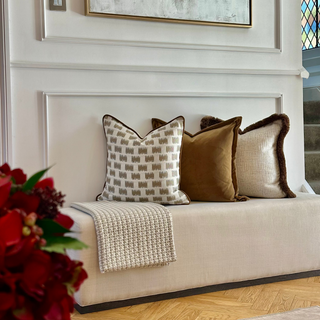 A styled entryway or lounge corner featuring a cream upholstered bench with three decorative throw pillows in beige, brown, and cream with dark trim, a cream knit throw blanket, and a large abstract framed painting on the wall above.