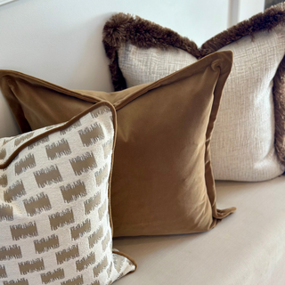 A styled collection of home decor throw pillows on a light beige sofa. The set includes a solid camel brown velvet pillow, a cream and taupe geometric patterned pillow with brown trim, and a cream textured pillow with a dark brown faux fur trim.