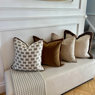 A styled entryway bench with five decorative throw pillows in neutral and earthy tones, including a patterned beige pillow, a brown velvet pillow, and textured cream pillows with dark trim, set against a white paneled wall with a framed abstract print.