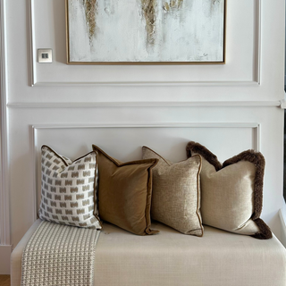 A framed abstract wall art piece with a gold-toned frame hangs on a white paneled wall above a beige upholstered bench with four decorative throw pillows.