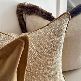 Close-up of decorative throw pillows on a sofa, including a front pillow in textured beige linen with brown piping, a brown velvet pillow, and a cream pillow with a dark brown faux-fur trim against a white wall.