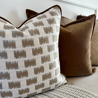 A set of decorative throw pillows arranged on a light-colored sofa. The front pillow features a cream and taupe ikat-style geometric pattern with dark brown piping, while the background pillows include a solid taupe velvet pillow, a textured beige linen pillow, and a cream pillow with a dark brown faux-fur trim.