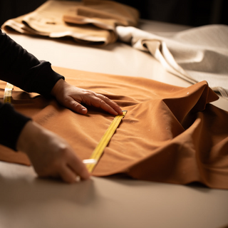 Person measuring brown fabric with a tape measure on a table.
