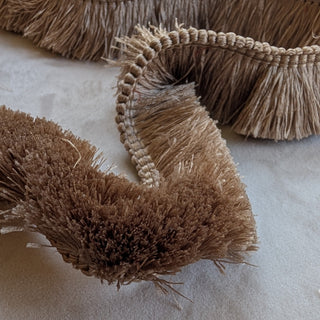A close-up of a decorative fringed trim or curtain edge in warm tan and brown tones, draped on a light neutral fabric background.