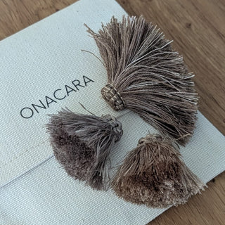 A pair of brown tassel earrings with a textured metallic cap, resting on a white canvas jewelry pouch printed with the brand name 'ONACARA', set on a wooden tabletop.